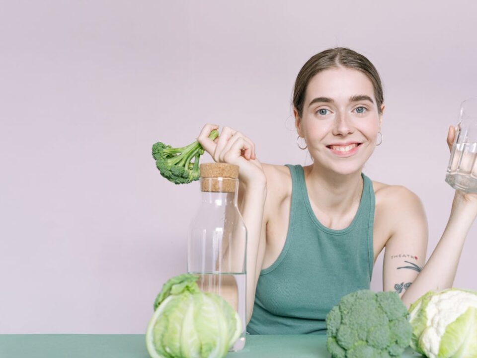 A woman in a green tank top holds a glass of water and a stalk of broccoli to illustrate Bellevue restaurant options that are good for your teeth.